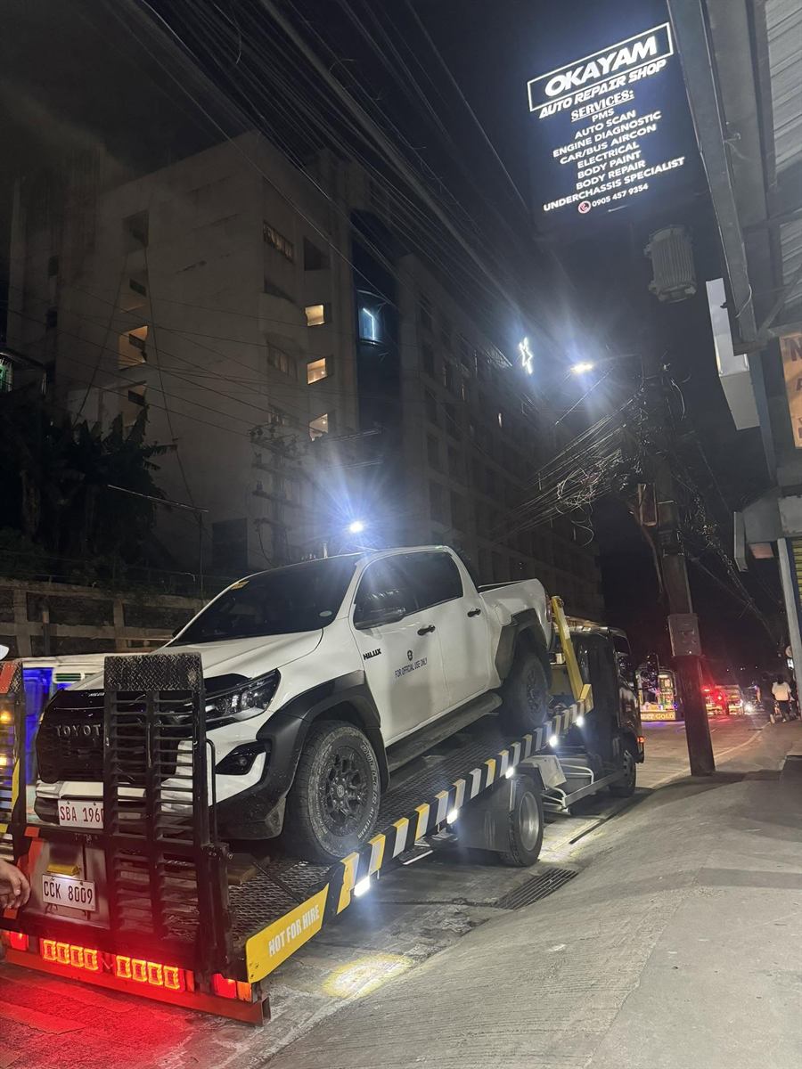 A white Toyota Hilux being winched onto the Okayam flatbed at night, the lit shop sign overhead