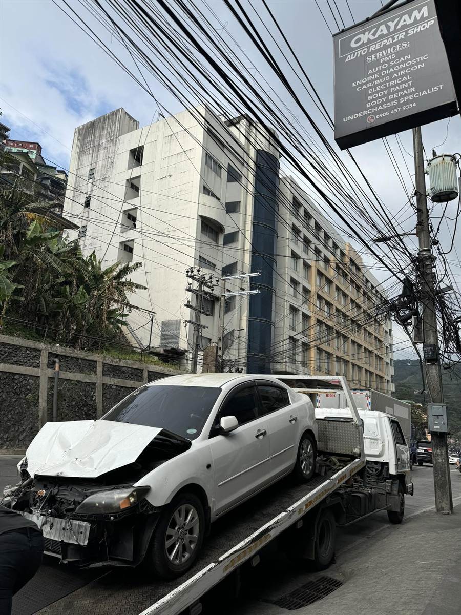 A white Mazda 3 with its front end crushed, loaded on the Okayam flatbed on Magsaysay Avenue, the shop sign overhead