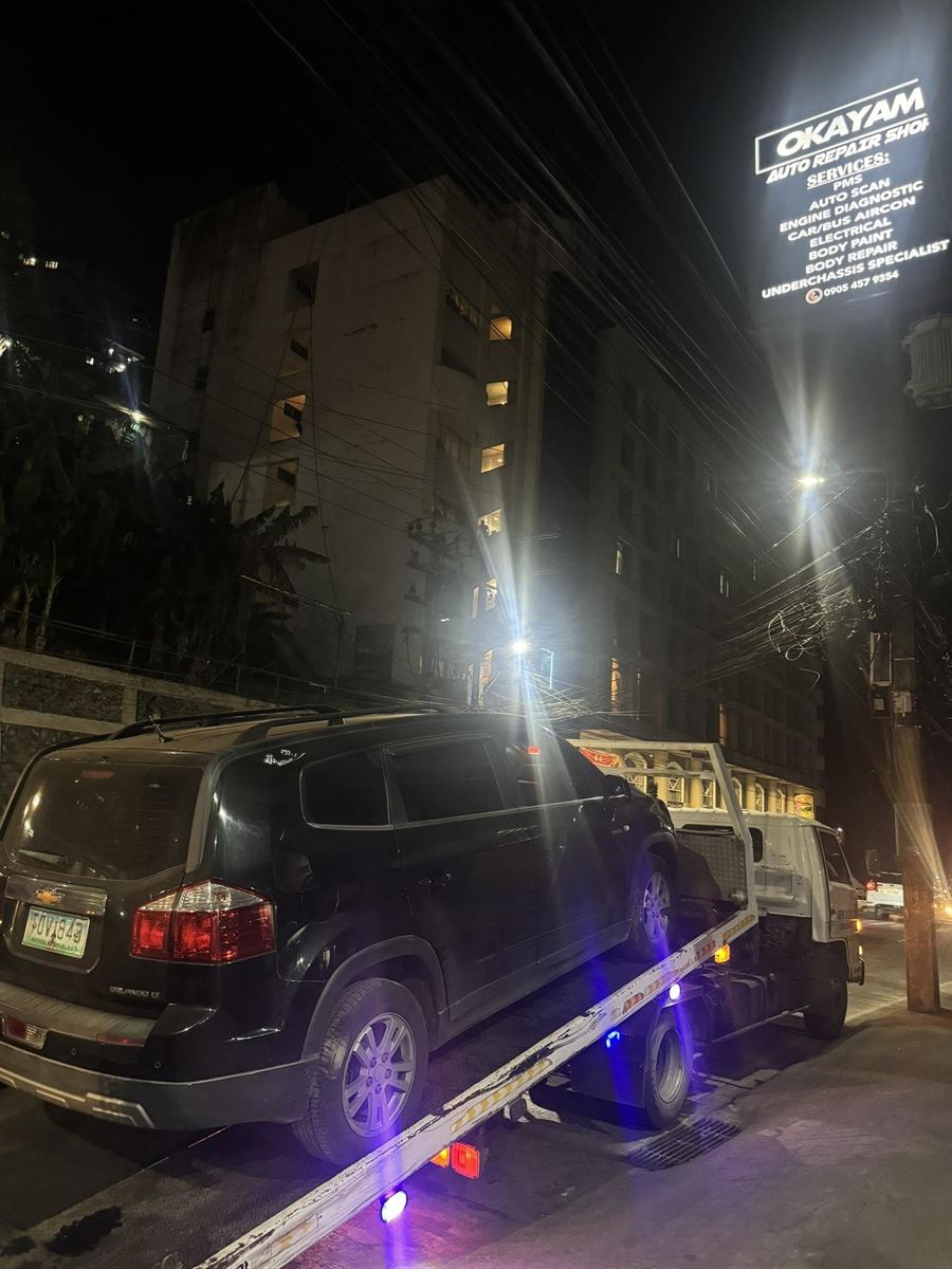 A black Chevrolet Orlando winched onto the Okayam flatbed at night under the lit shop sign